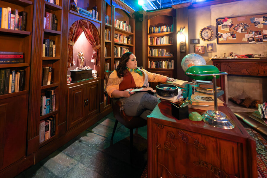 person sits in chair with largen wooden bookcase behind and wooden desk in front with globe and other items
